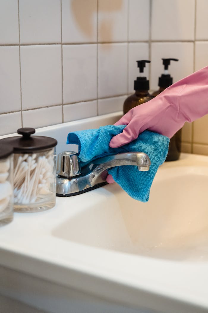 gallery-01 A gloved hand cleans a bathroom sink faucet with a blue cloth for hygiene.
