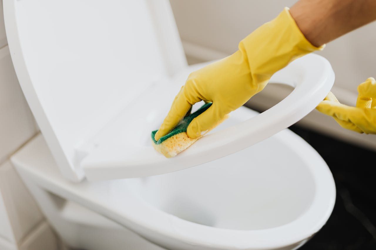 services-01 A person using yellow gloves and a sponge to clean a toilet seat indoors.